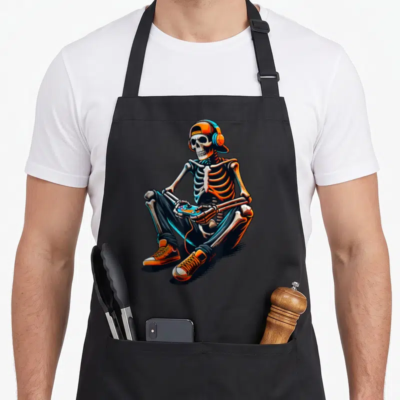 Skeleton gamer video gaming halloween apron - Cartoon skeleton in orange and blue sitting cross-legged with a game — shown on a black cotton canvas apron against a white background