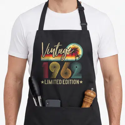 Edition 63rd birthday apron - Vintage 1962 LIMITED EDITION text over retro sunset with palm trees in beige red orange — shown on a black cotton canvas apron against a white background