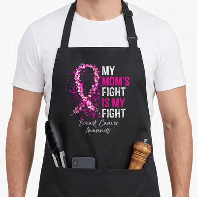 Fight breast cancer awareness aprons - Pink ribbon made of small hearts next to stacked text reading My Mom's Fight is — shown on a black cotton canvas apron against a white background