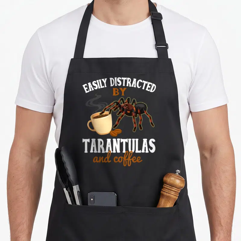Tarantula coffee apron - Cartoon tarantula with brown legs leaning over cream cup next to text easily distracted — shown on a black cotton canvas apron against a white background