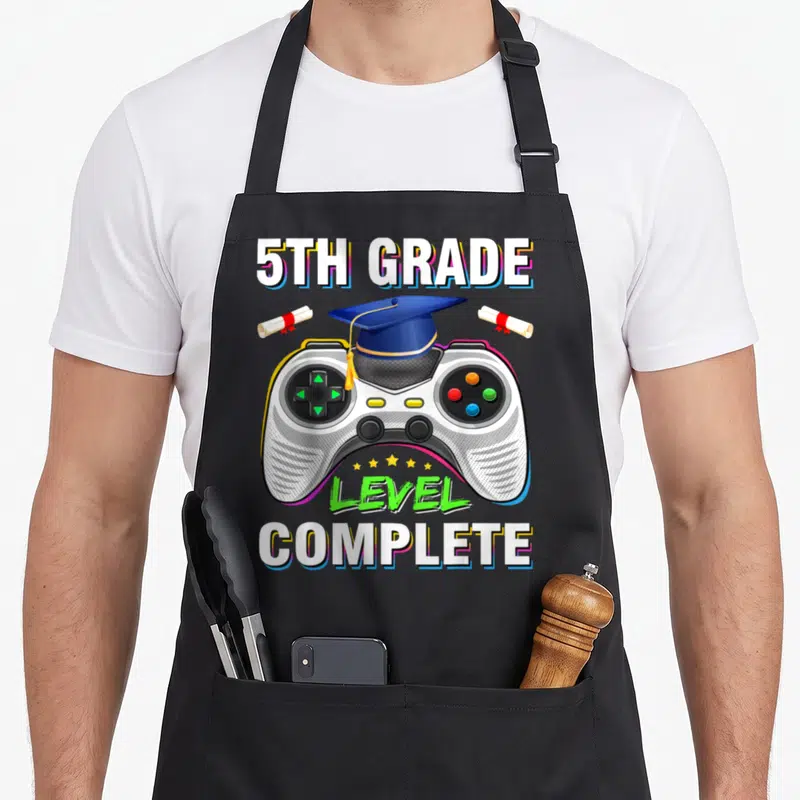 5th gaming apron - Game controller with graduation cap, two diplomas, and 5th Grade Level Complete text — shown on a black cotton canvas apron against a white background