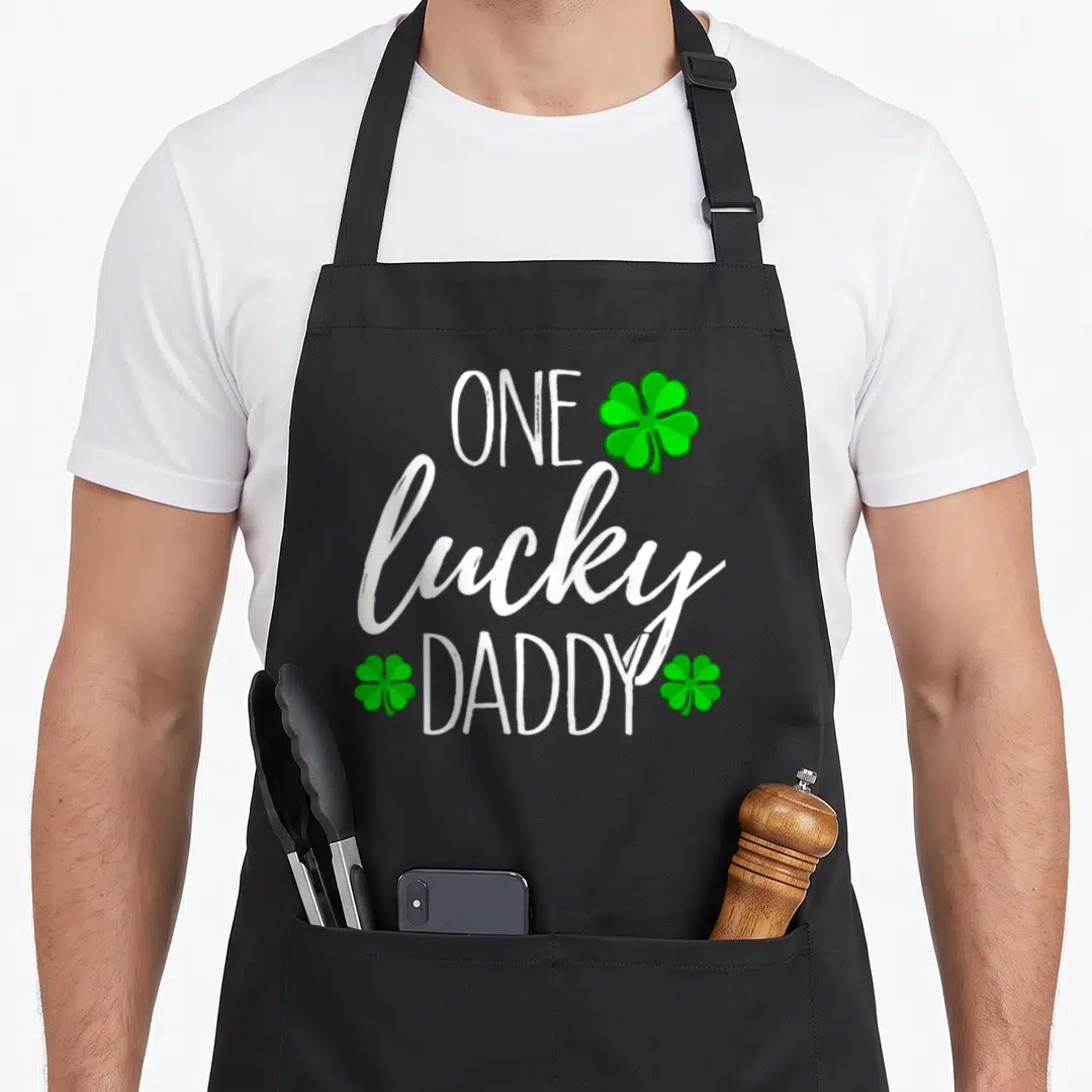 Cute lucky st patrick day aprons - ONE lucky DADDY white text with three green shamrocks top right, bottom left, bottom — shown on a black cotton canvas apron against a white background