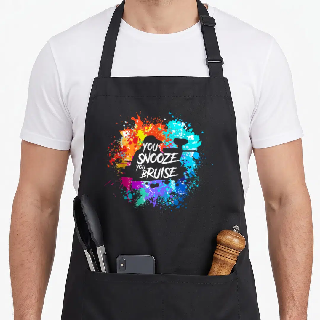 Funny snooze paintball apron - Distressed white text You Snooze You Bruise over grey silhouette and colorful paint — shown on a black cotton canvas apron against a white background