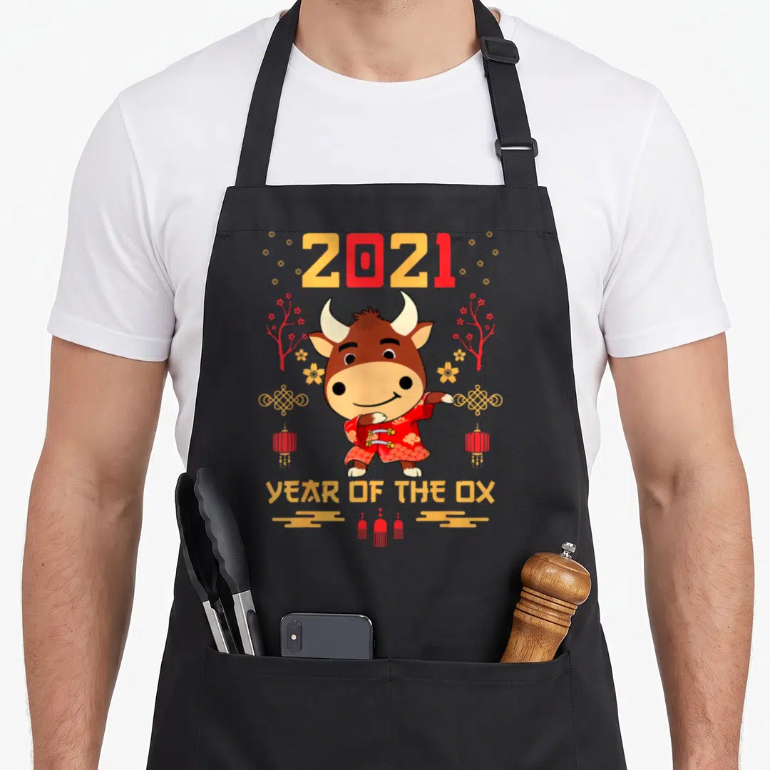 Clothe year of the ox apron - Cartoon brown ox in red robe with 2021 text, lanterns, and knots — shown on a black cotton canvas apron against a white background