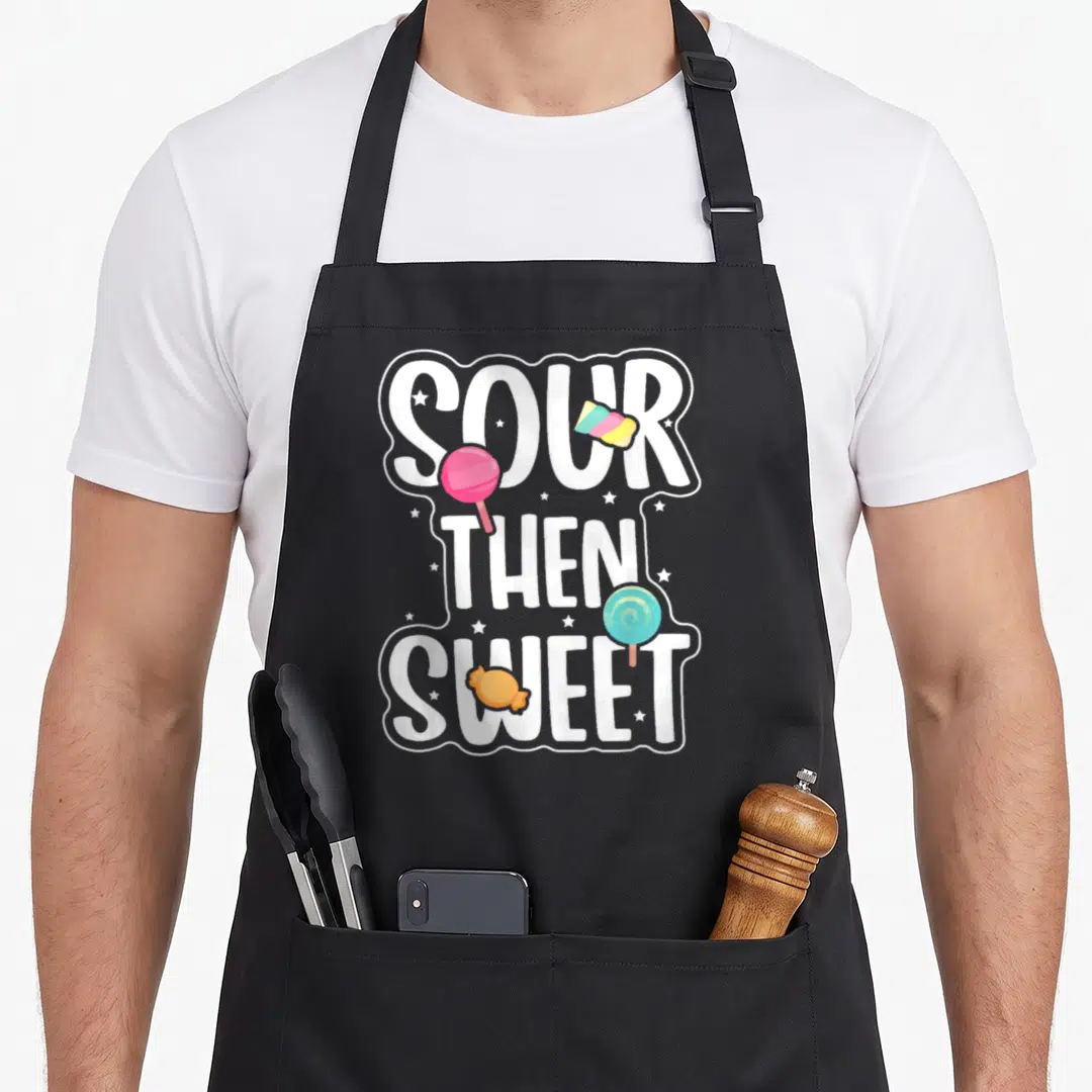 Patch candy aprons - SOUR THEN SWEET text with pink blue yellow lollipops and wrapped candies — shown on a black cotton canvas apron against a white background
