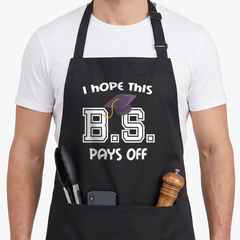Bachelor diploma apron - I Hope This B.S. Pays Off text with purple graduation cap and white block letters — shown on a black cotton canvas apron against a white background