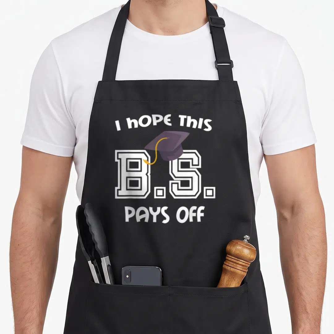 Bachelor diploma apron - I Hope This B.S. Pays Off text with purple graduation cap and white block letters — shown on a black cotton canvas apron against a white background