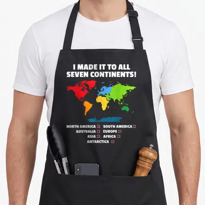 Seven traveler apron - World map in red blue green yellow orange with checked continent list and bold white text — shown on a black cotton canvas apron against a white background