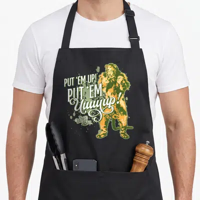 Wizard lion apron - Cowardly Lion in orange and green with stacked PUT EM UP text and Wizard of Oz logo — shown on a black cotton canvas apron against a white background