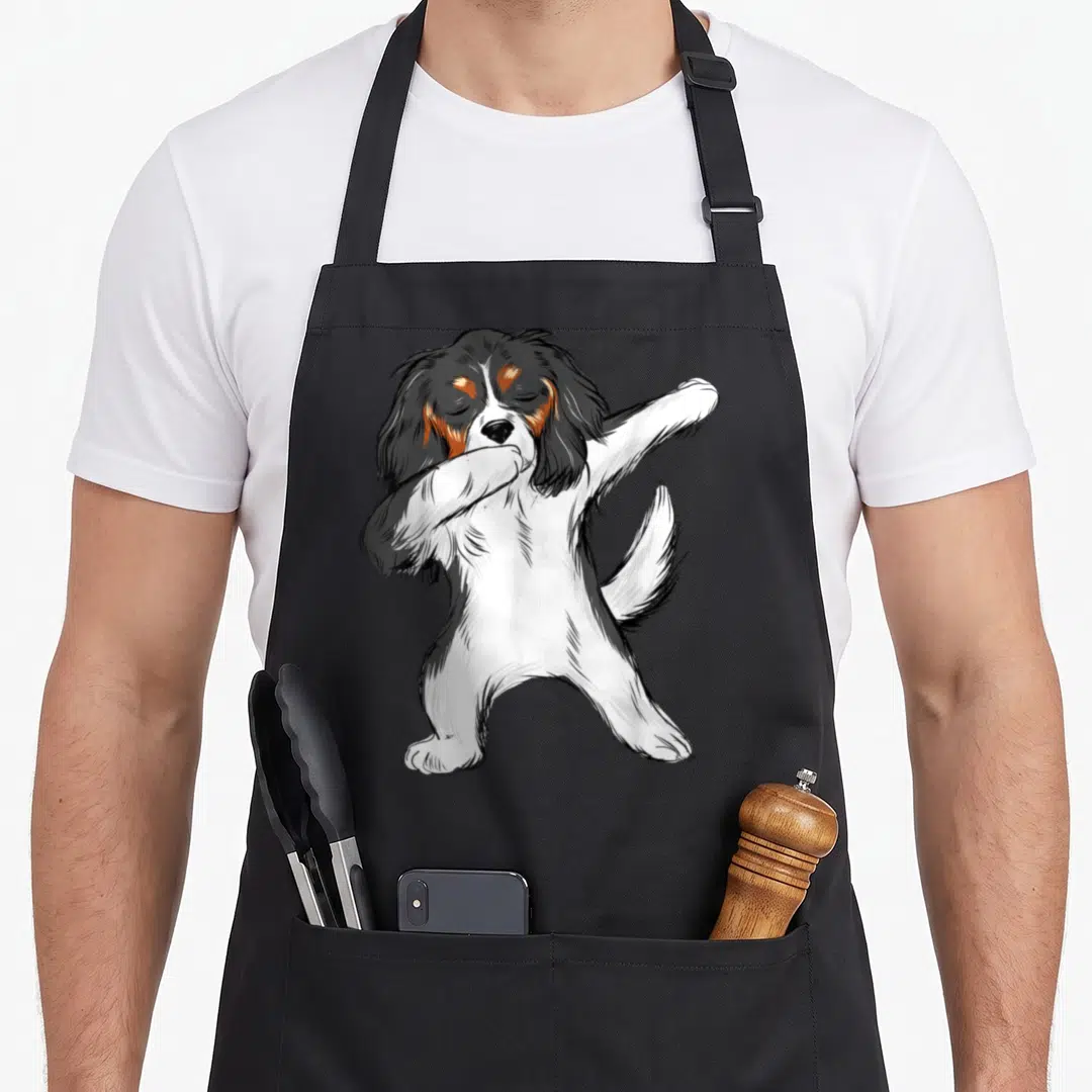 Dabbing king apron - Cartoon spaniel standing on two legs dabbing in white, black, and brown — shown on a black cotton canvas apron against a white background
