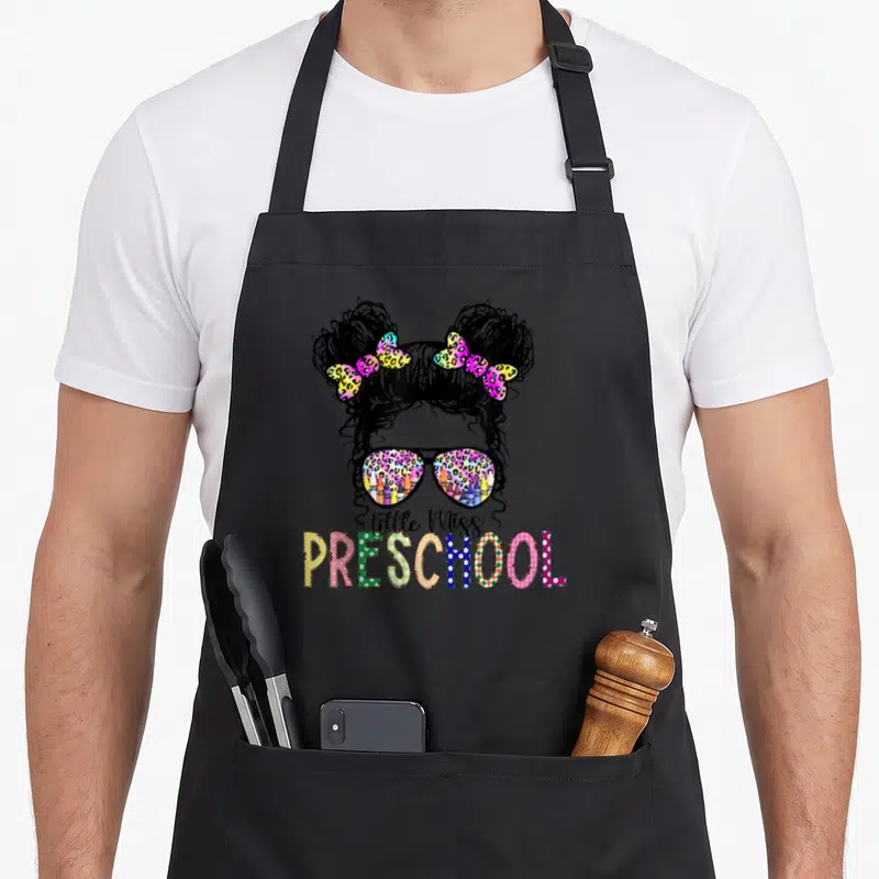 Miss preschool tie-dye apron - Cartoon girl head with messy buns, leopard sunglasses, and Little Miss PRESCHOOL text — shown on a black cotton canvas apron against a white background