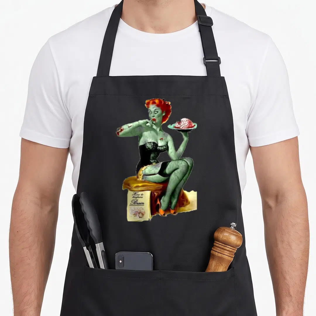 Zombie pin up apron - Green zombie woman on yellow stool holding brain plate with How to prepare Brain book — shown on a black cotton canvas apron against a white background