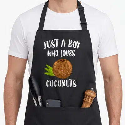 Funny coconut apron - Just A Boy Who Loves Coconuts text above and below a smiling cartoon coconut with green leaves — shown on a black cotton canvas apron against a white background