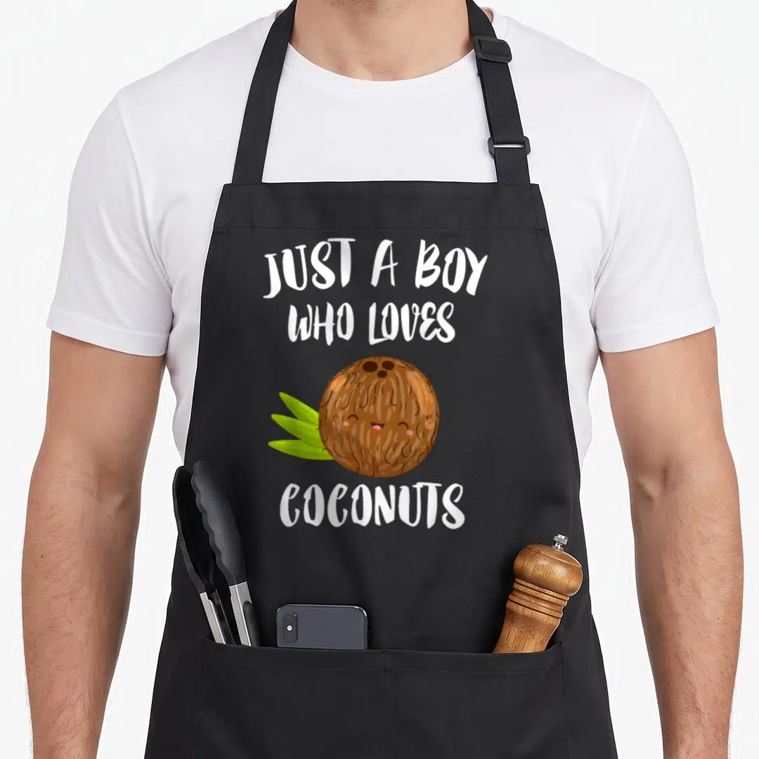 Funny coconut apron - Just A Boy Who Loves Coconuts text above and below a smiling cartoon coconut with green leaves — shown on a black cotton canvas apron against a white background
