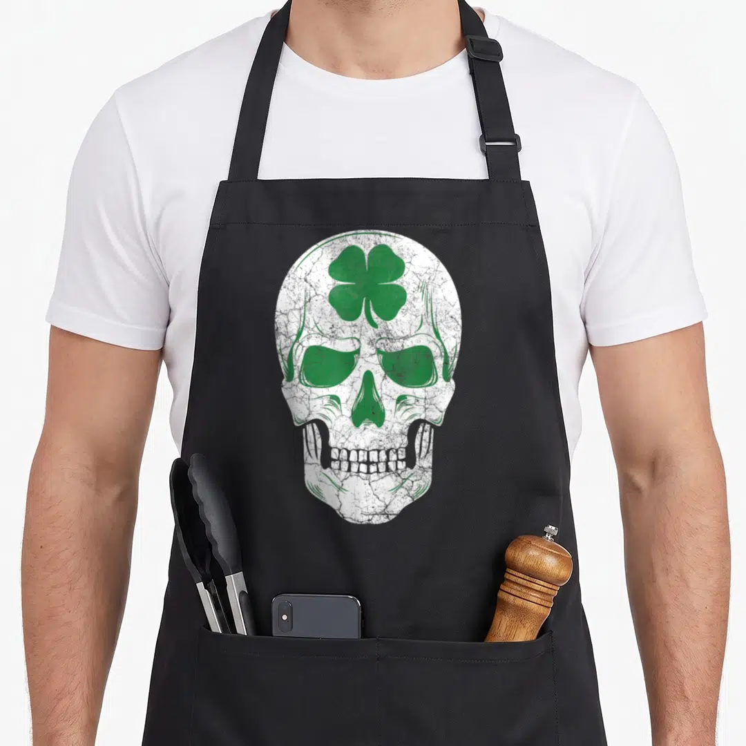 Shamrock ireland apron - White distressed skull with green four-leaf clover on forehead — shown on a black cotton canvas apron against a white background