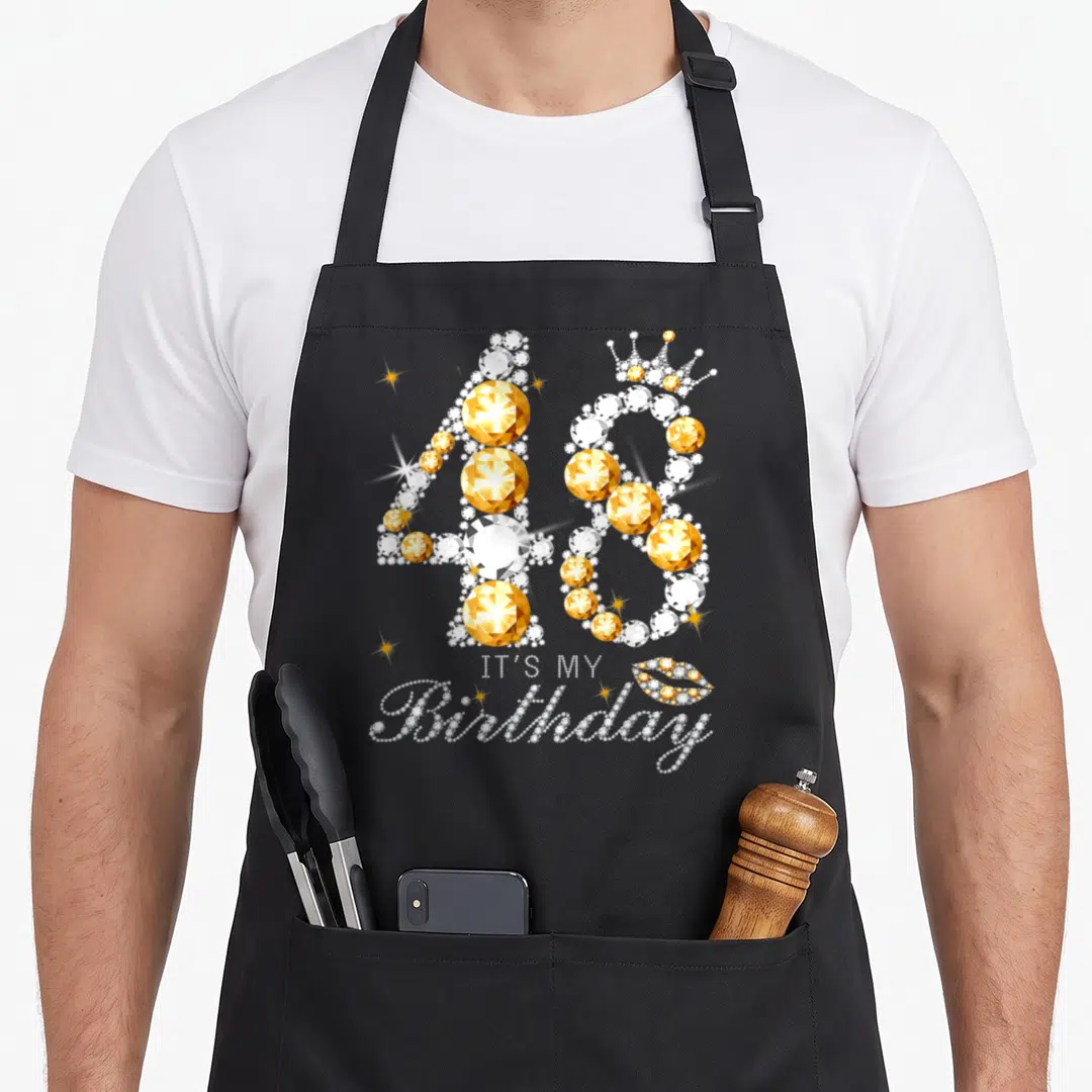 48th queen of baking apron - Jeweled number 48 with crown and IT'S MY Birthday text in white and gold — shown on a black cotton canvas apron against a white background