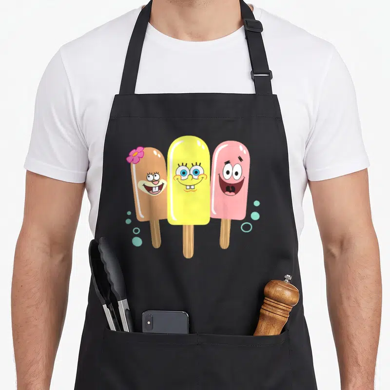 Sandy ice cream apron - Three cartoon popsicles in brown, yellow, and pink with a flower and blue bubbles — shown on a black cotton canvas apron against a white background