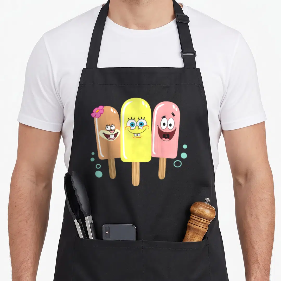 Sandy ice cream apron - Three cartoon popsicles in brown, yellow, and pink with a flower and blue bubbles — shown on a black cotton canvas apron against a white background