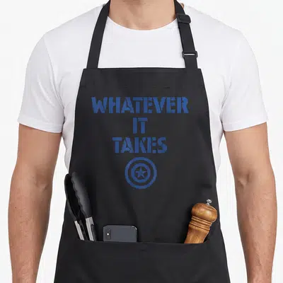 Cute endgame whatever apron - Distressed blue text reading Whatever It Takes above a circular star shield symbol — shown on a black cotton canvas apron against a white background