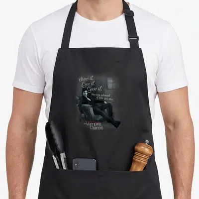 Vampire big brother apron - Man in armchair with Own it Live it Love it quote and Vampire Diaries logo — shown on a black cotton canvas apron against a white background