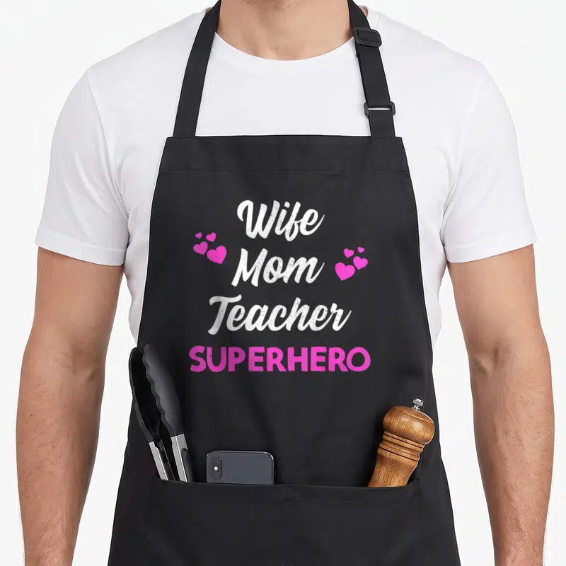 Mother best teacher apron - Stacked text Wife Mom Teacher SUPERHERO with pink heart clusters flanking the word Mom — shown on a black cotton canvas apron against a white background