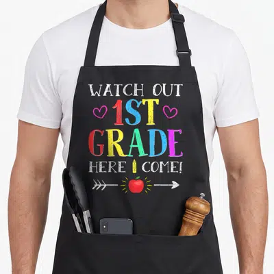 Grade novelty apron - Colorful text WATCH OUT 1ST GRADE HERE I COME with pencil and apple graphics — shown on a black cotton canvas apron against a white background