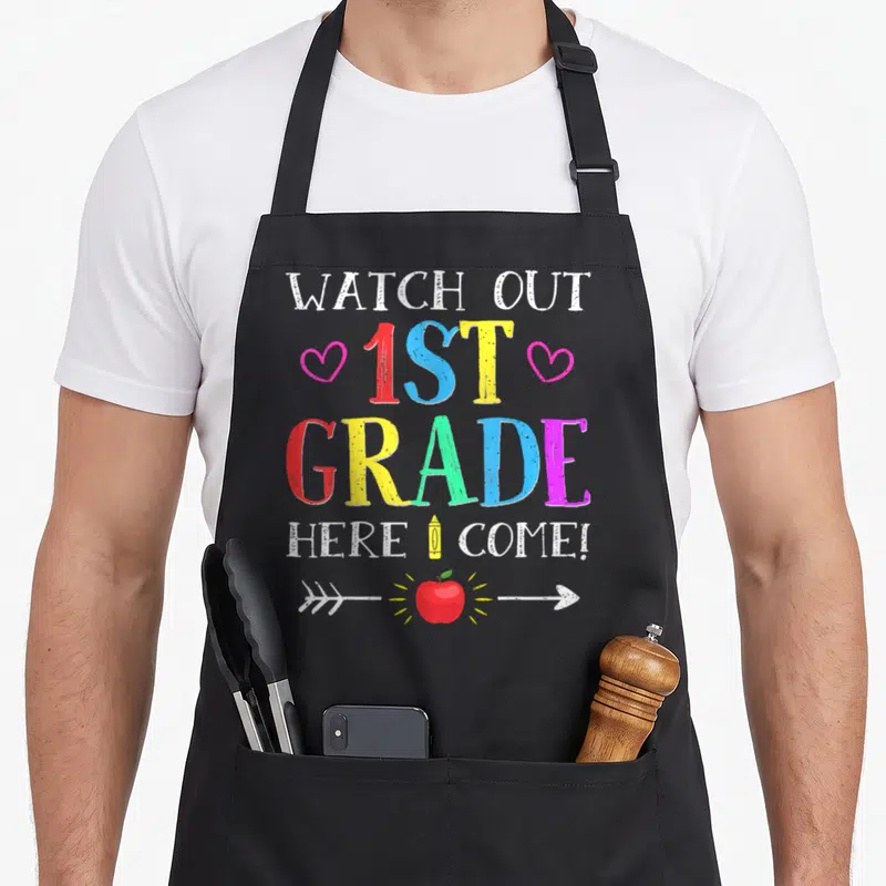 Grade novelty apron - Colorful text WATCH OUT 1ST GRADE HERE I COME with pencil and apple graphics — shown on a black cotton canvas apron against a white background