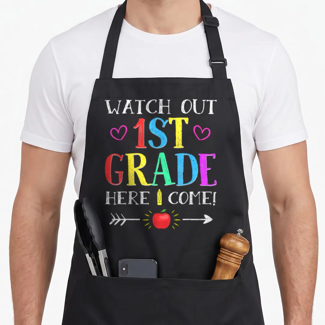 Grade novelty apron - Colorful text WATCH OUT 1ST GRADE HERE I COME with pencil and apple graphics — shown on a black cotton canvas apron against a white background