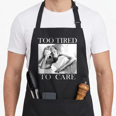 Tired marilyn monroe apron - Grayscale photo of woman in bed with text too tired to care — shown on a black cotton canvas apron against a white background