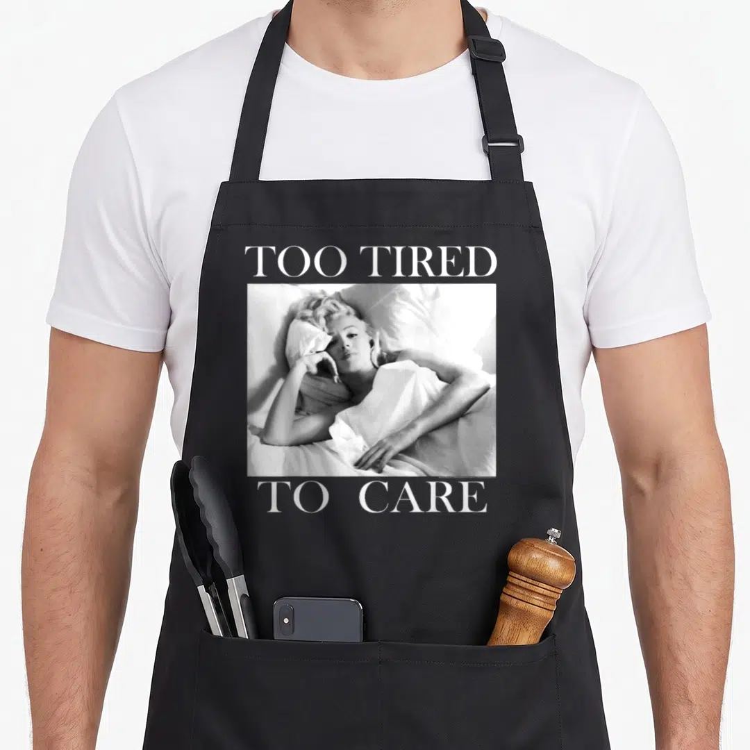 Tired marilyn monroe apron - Grayscale photo of woman in bed with text too tired to care — shown on a black cotton canvas apron against a white background