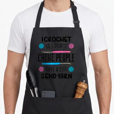 Crocheter knitting apron - I CROCHET SO I DON'T CHOKE PEOPLE text with pink blue hooks and yarn balls — shown on a black cotton canvas apron against a white background