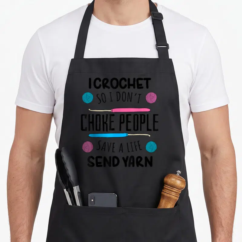 Crocheter knitting apron - I CROCHET SO I DON'T CHOKE PEOPLE text with pink blue hooks and yarn balls — shown on a black cotton canvas apron against a white background