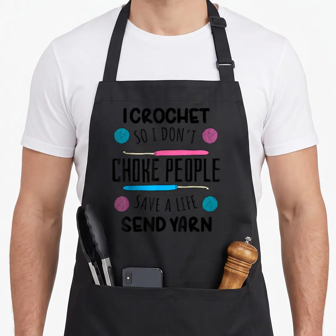 Crocheter knitting apron - I CROCHET SO I DON'T CHOKE PEOPLE text with pink blue hooks and yarn balls — shown on a black cotton canvas apron against a white background