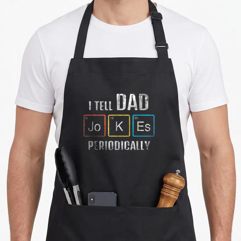 Funny periodically dad joke apron - I TELL DAD Jo K Es PERIODICALLY text in red yellow blue squares — shown on a black cotton canvas apron against a white background