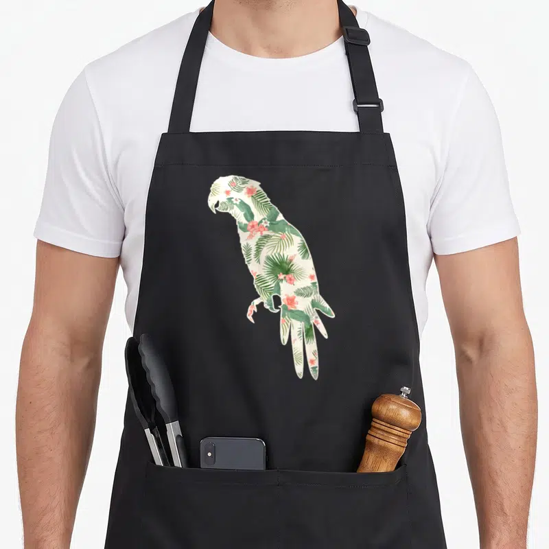Parrot the floral apron - Parrot silhouette filled with green tropical leaves and pink flowers on cream background — shown on a black cotton canvas apron against a white background