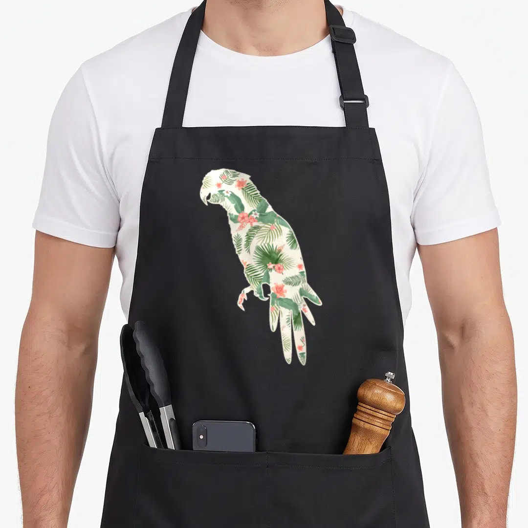 Parrot the floral apron - Parrot silhouette filled with green tropical leaves and pink flowers on cream background — shown on a black cotton canvas apron against a white background