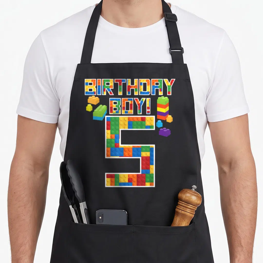 5th building apron - Colorful brick letters spelling BIRTHDAY BOY! 5 with scattered blocks — shown on a black cotton canvas apron against a white background