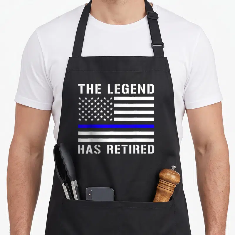 Legend line apron - White and blue distressed flag with The Legend Has Retired text and thin blue line stripe — shown on a black cotton canvas apron against a white background