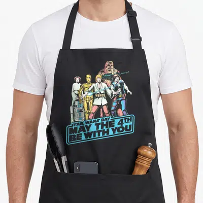 Shot fourth apron - Star Wars characters standing behind blue banner reading May The 4th Be With You — shown on a black cotton canvas apron against a white background