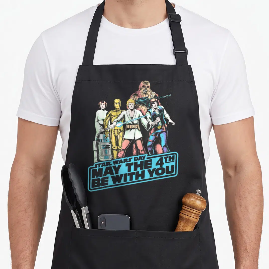 Shot fourth apron - Star Wars characters standing behind blue banner reading May The 4th Be With You — shown on a black cotton canvas apron against a white background