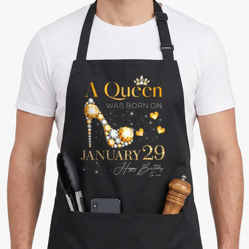 January 29th birthday apron - Gold high heel shoe formed by gems with crown and text A Queen Was Born on January 29 — shown on a black cotton canvas apron against a white background