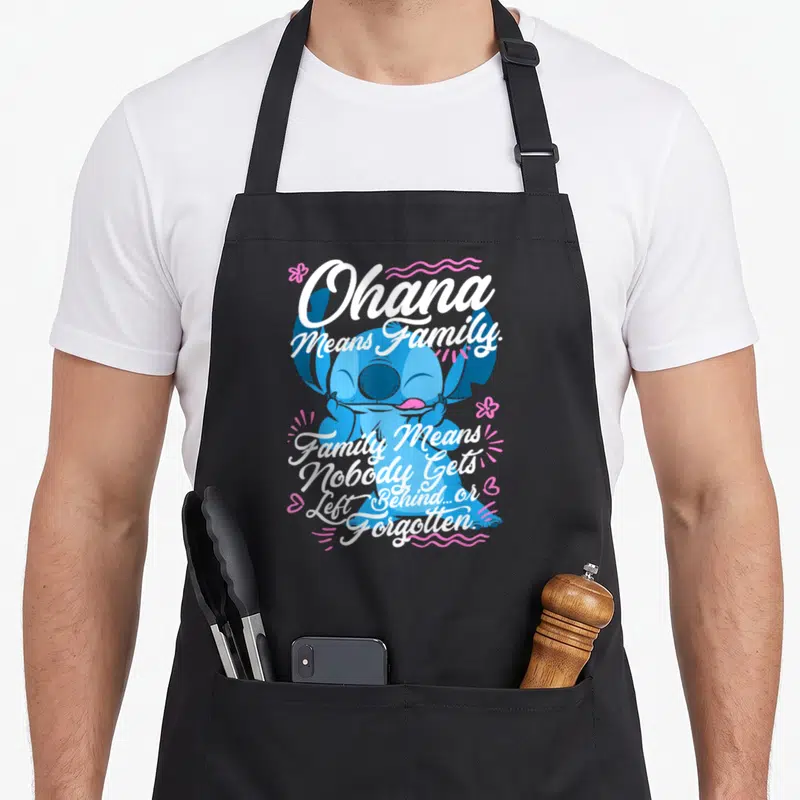 Family stitch apron - Blue cartoon character with white Ohana Means Family text and pink floral accents — shown on a black cotton canvas apron against a white background