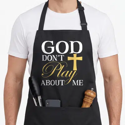 Play faith aprons - Stacked white and gold text reading God Don't Play About Me with a gold cross overlay — shown on a black cotton canvas apron against a white background