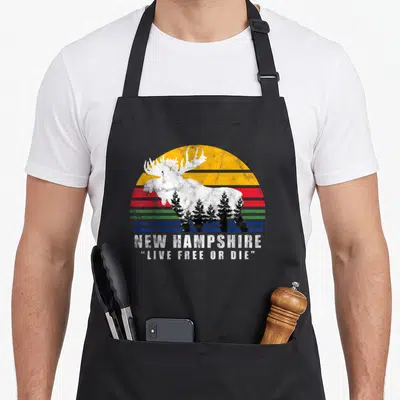 Die apron or apron - White moose with pine trees over striped sun, NEW HAMPSHIRE LIVE FREE OR DIE text — shown on a black cotton canvas apron against a white background