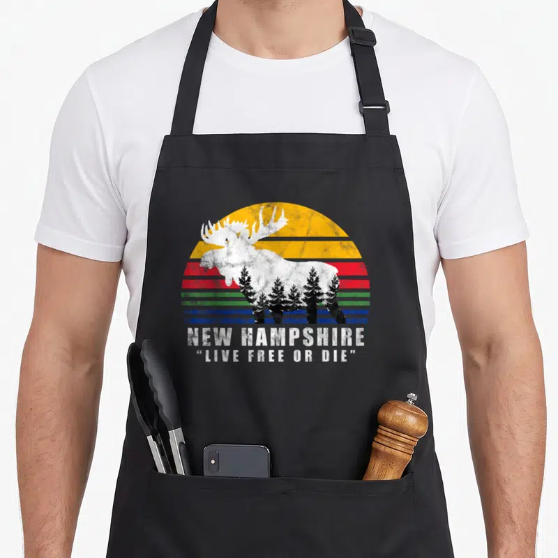 Die apron or apron - White moose with pine trees over striped sun, NEW HAMPSHIRE LIVE FREE OR DIE text — shown on a black cotton canvas apron against a white background