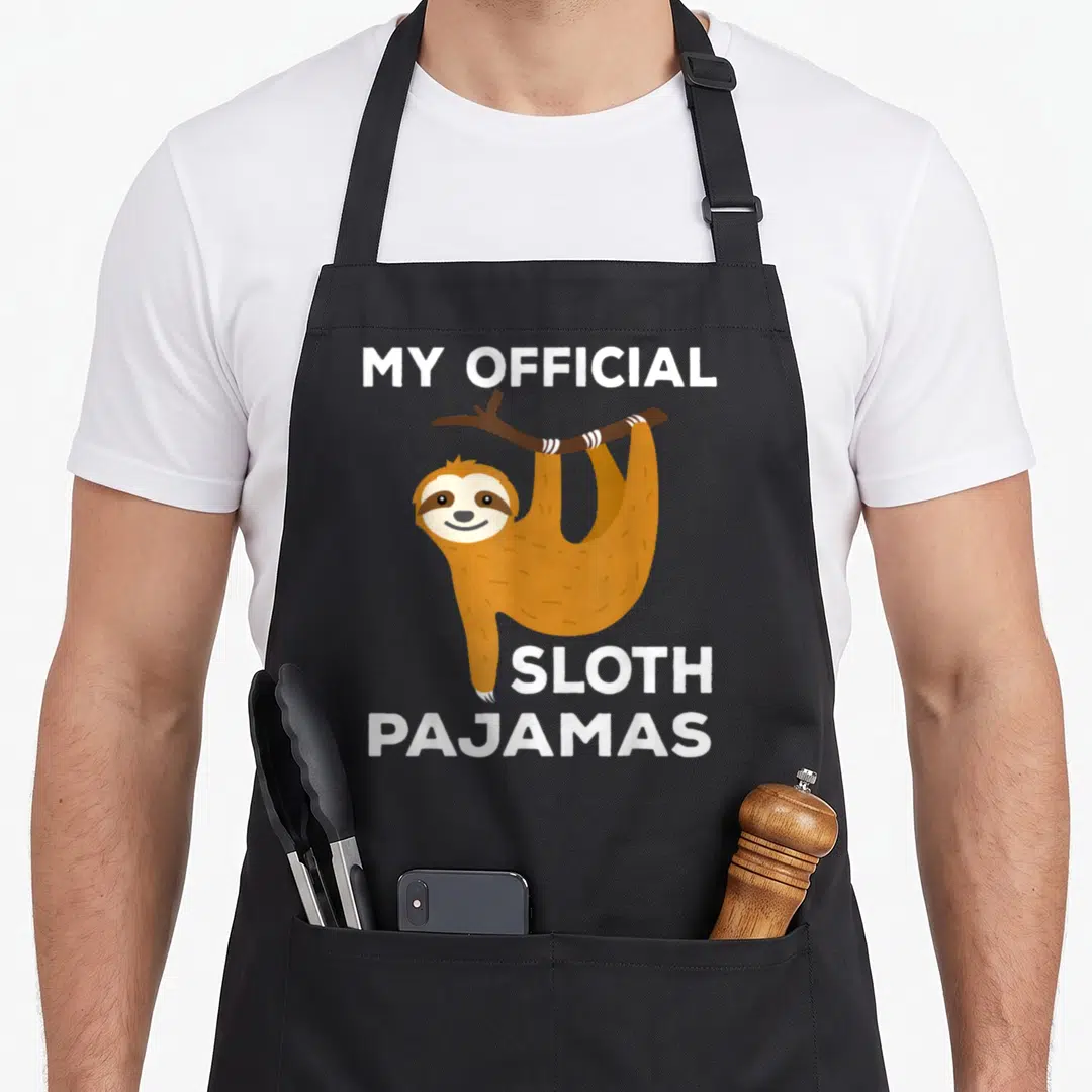 Everyday summer aprons - Cartoon sloth hanging from brown branch with white text MY OFFICIAL SLOTH PAJAMAS — shown on a black cotton canvas apron against a white background