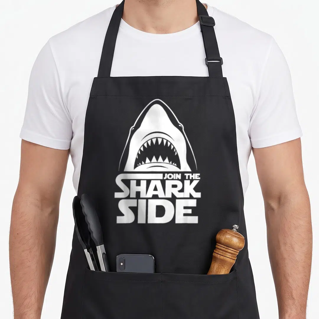 Join shark lover apron - White shark head above stacked text JOIN THE SHARK SIDE — shown on a black cotton canvas apron against a white background
