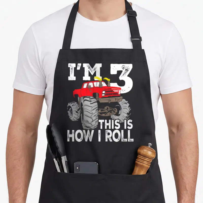 3rd truck apron - Red monster truck with white text I'M 3 THIS IS HOW I ROLL in cartoon style — shown on a black cotton canvas apron against a white background