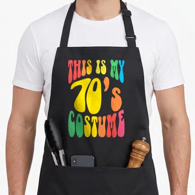 Costume hippie apron - Stacked psychedelic text reading THIS IS MY 70S COSTUME in rainbow colors — shown on a black cotton canvas apron against a white background