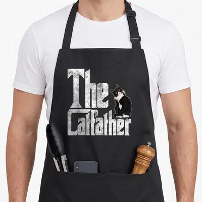 Catfather daddy aprons - White distressed text The Catfather beside a sitting black and white tuxedo cat — shown on a black cotton canvas apron against a white background